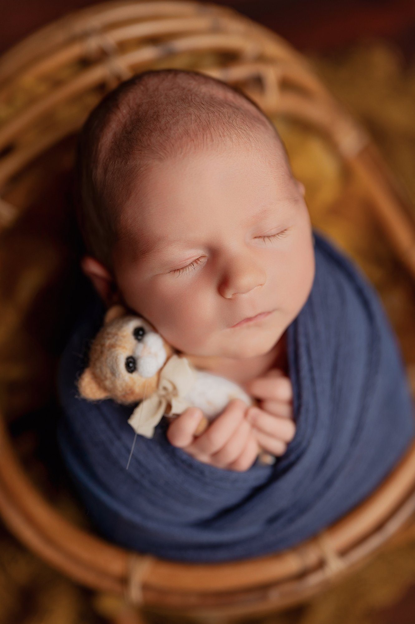 Sleeping newborn baby swaddled in navy blue wrap holding a tiny teddy bear, photographed from above in basket