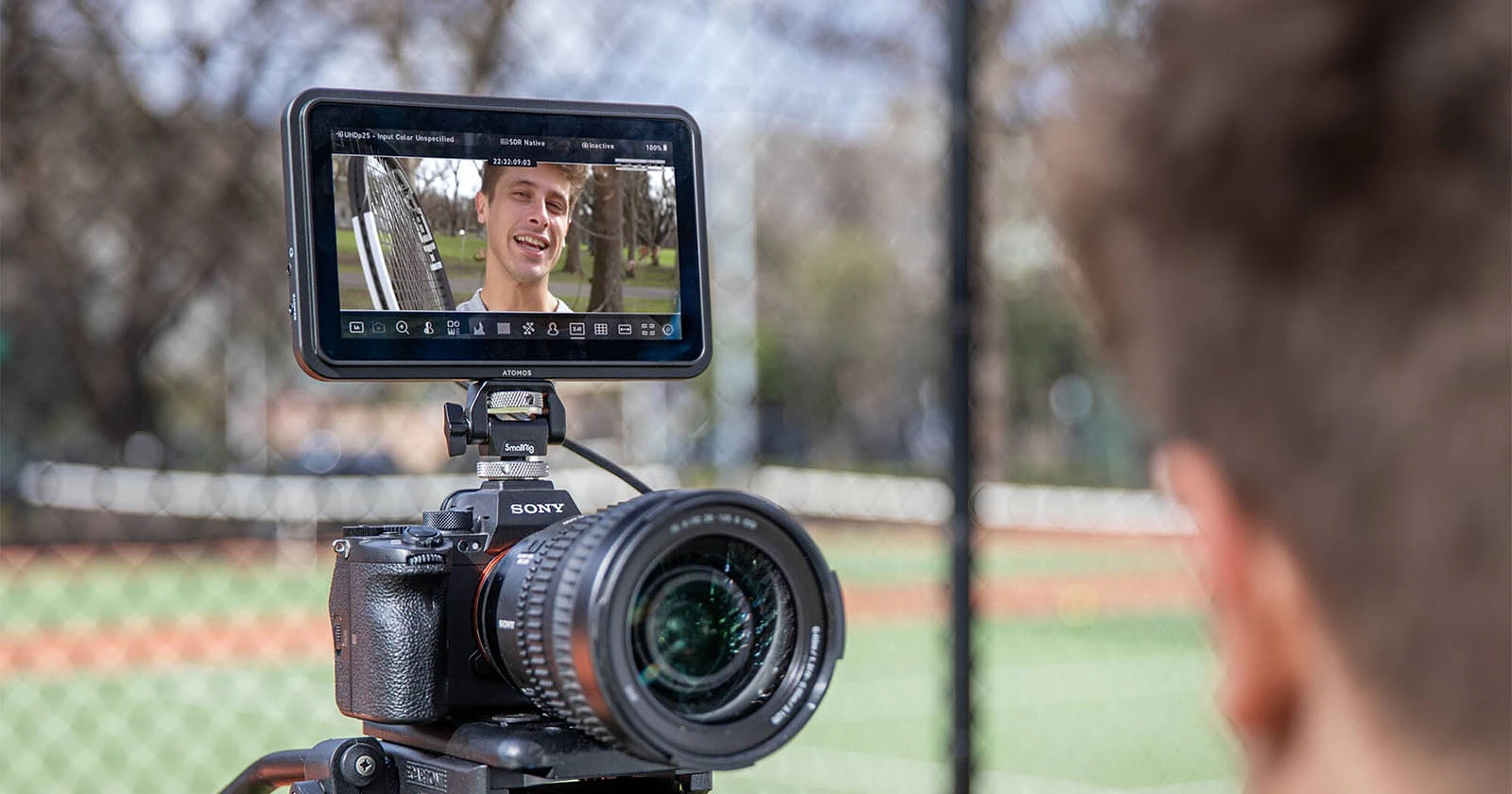 A camera on a tripod with an external monitor attached, showing a man holding a tennis racket, viewed from behind the shoulder of a person.