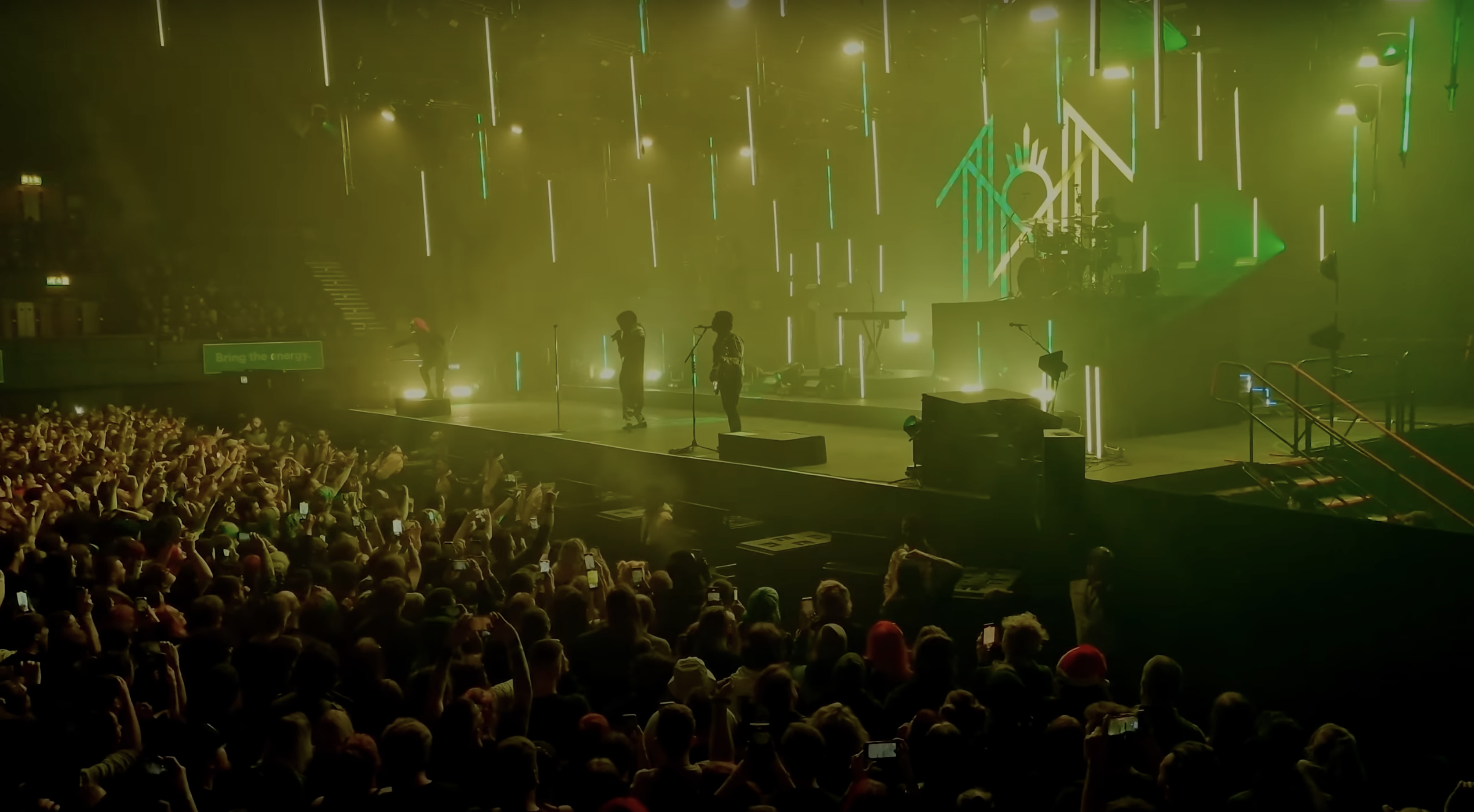 A video frame of Sleep Token performing Vore at Wembley. Facing the stage from the right, a large crowd cheers their performace, illuminated by the iconic green and yellow lights associated with the song. iii points into the crowd on the left as Vessel and iv sing in middle frame, and ii plays drums on his pedestal to the right of iv.