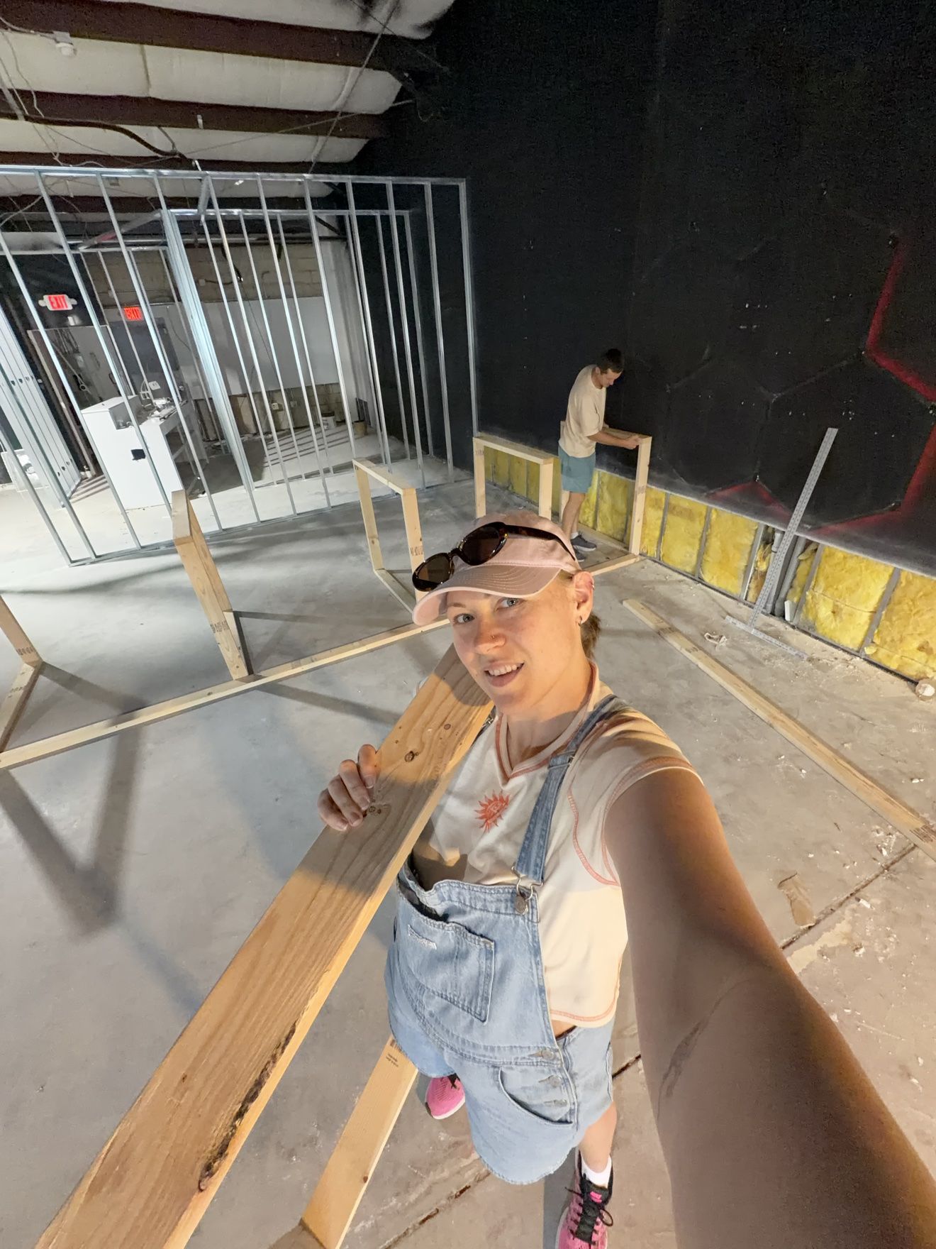 Masha carrying a long wooden board on her shoulder while Aleh builds a frame in the background