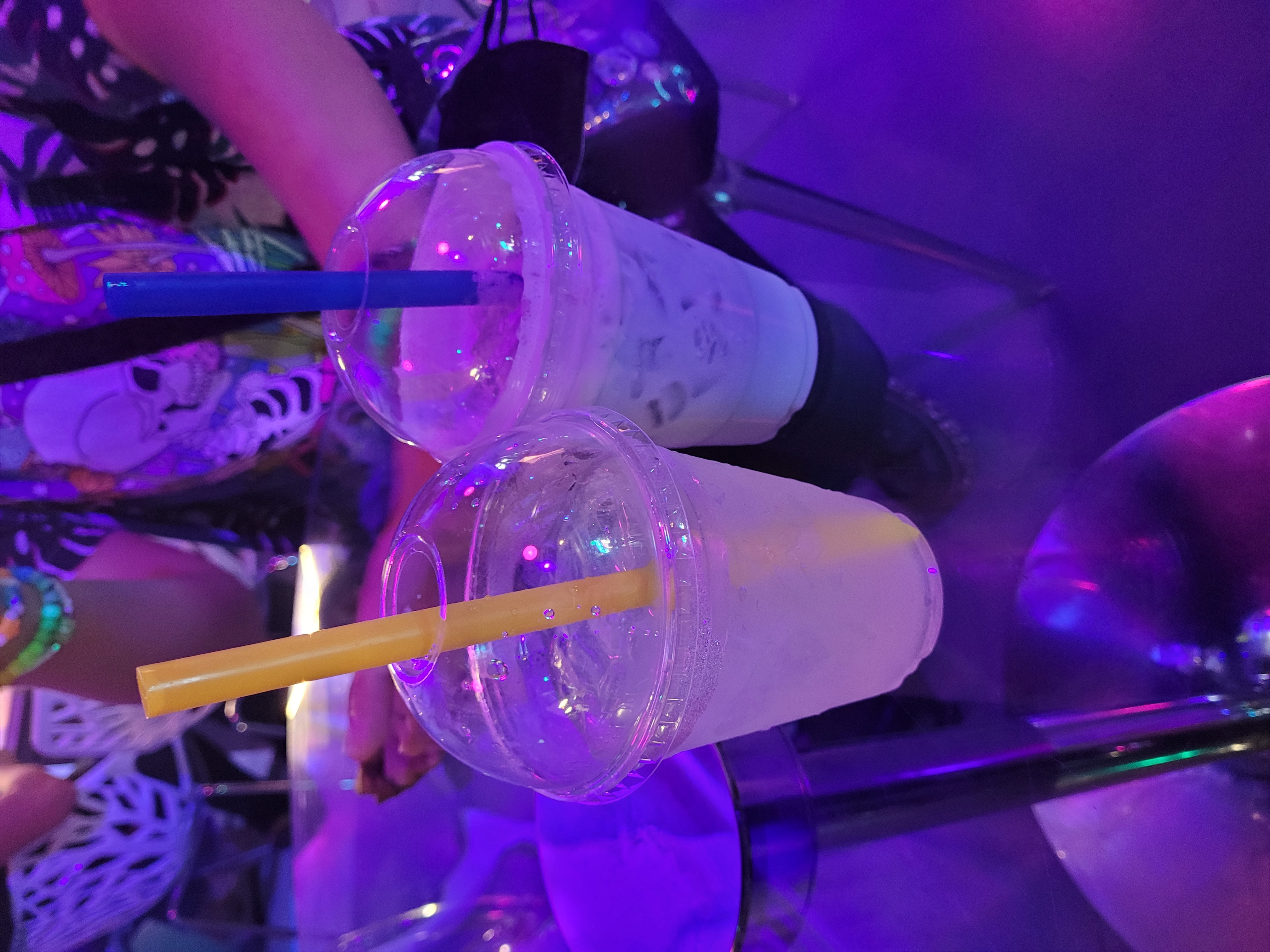 drinks served at the cafe. one is the sea tea, a marshmallow butterfly pea tea with vanilla seasalt cold foam. it is light blue, opaque, filled with ice and served in a transparent plastic cup with a dome top and a wide blue straw to match the blue color of the drink. the other drink is the reigen supreme, based off of reigen arataka from mob psycho 100 (notably not an undertale character. this is due in part due to at the time recent events, wherein on a twitter poll determining the ultimate tumblr sexyman, sans was pitted against reigen. said poll gained much notoriety during it's run and concluded mere hours prior to the queens death in september and in close proximity to when the cafe started, hence why it was notable at the time. it was also served due to the cafe being out of most everything else at the time we attended, and october tenth being reigen's stated birthday). it is a sparkling lemonade, semi-translucent and a very light yellow in color, again served with ice in a plastic cup with a dome lid and a wide straw, it being yellow to match the drink. the drinks sit atop a glass table, and the drinks and surrounding area are lit under the purple lights of the cafe.