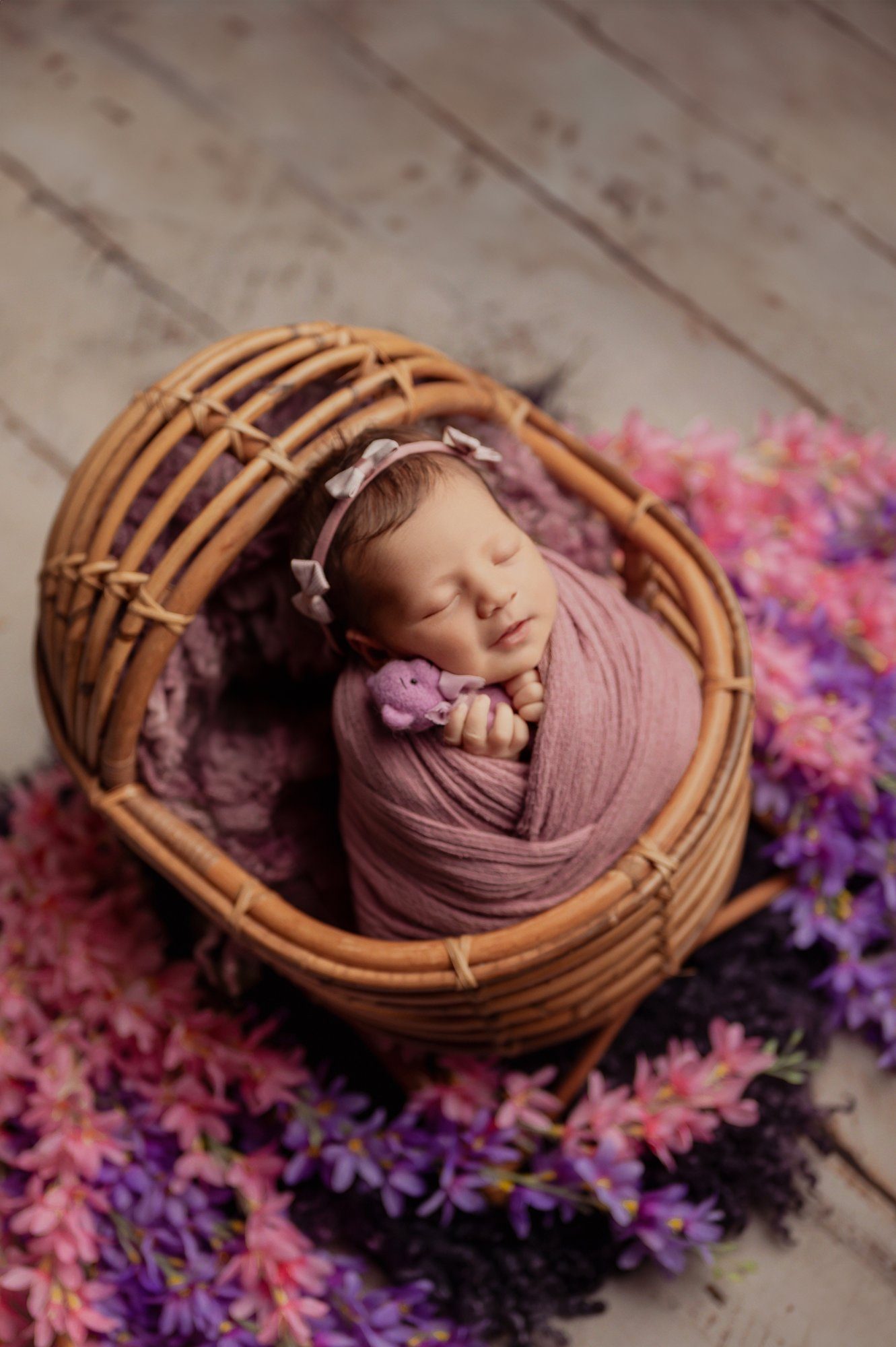Newborn baby sleeping peacefully in woven basket wrapped in mauve fabric surrounded by pink and purple dried flowers
