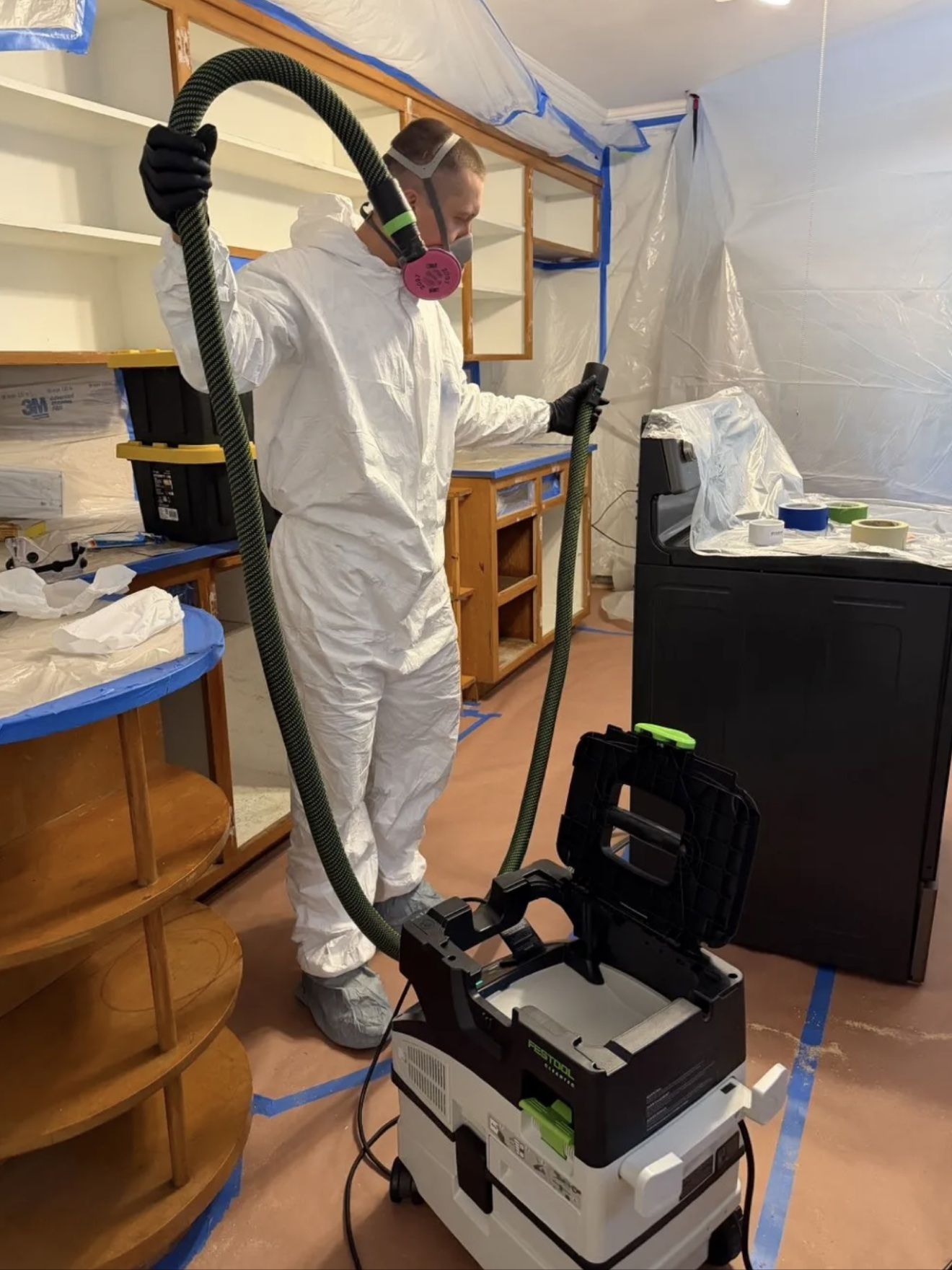 Aleh in a white protective suit with a dust extractor in a covered kitchen