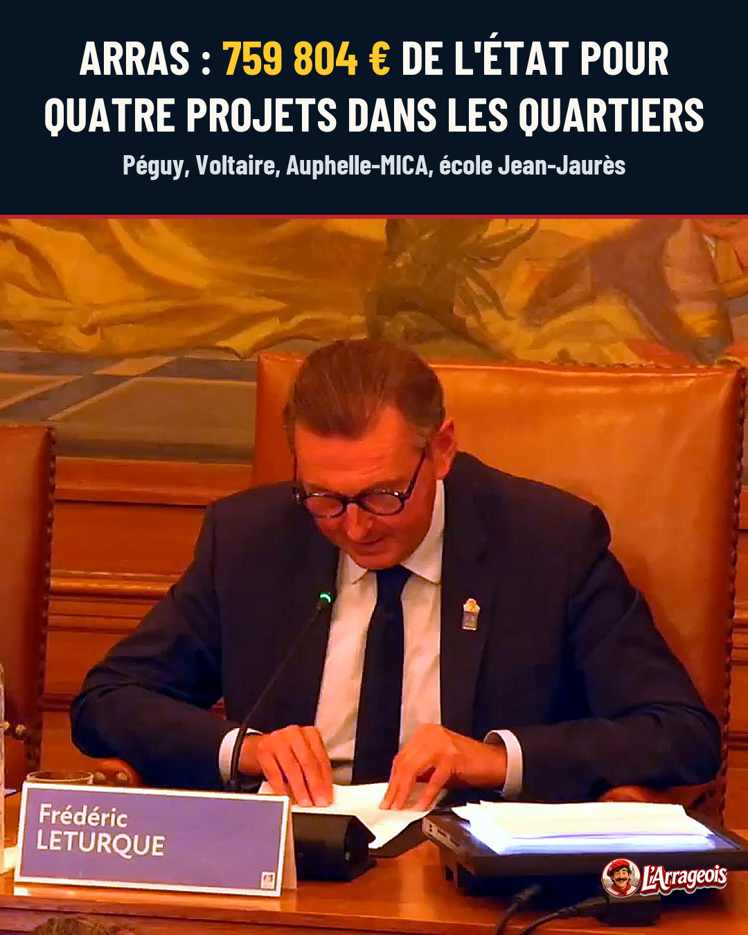 Un homme en costume sombre et cravate, portant des lunettes, est assis à une table avec un micro et un panneau nommable, regardant des documents.