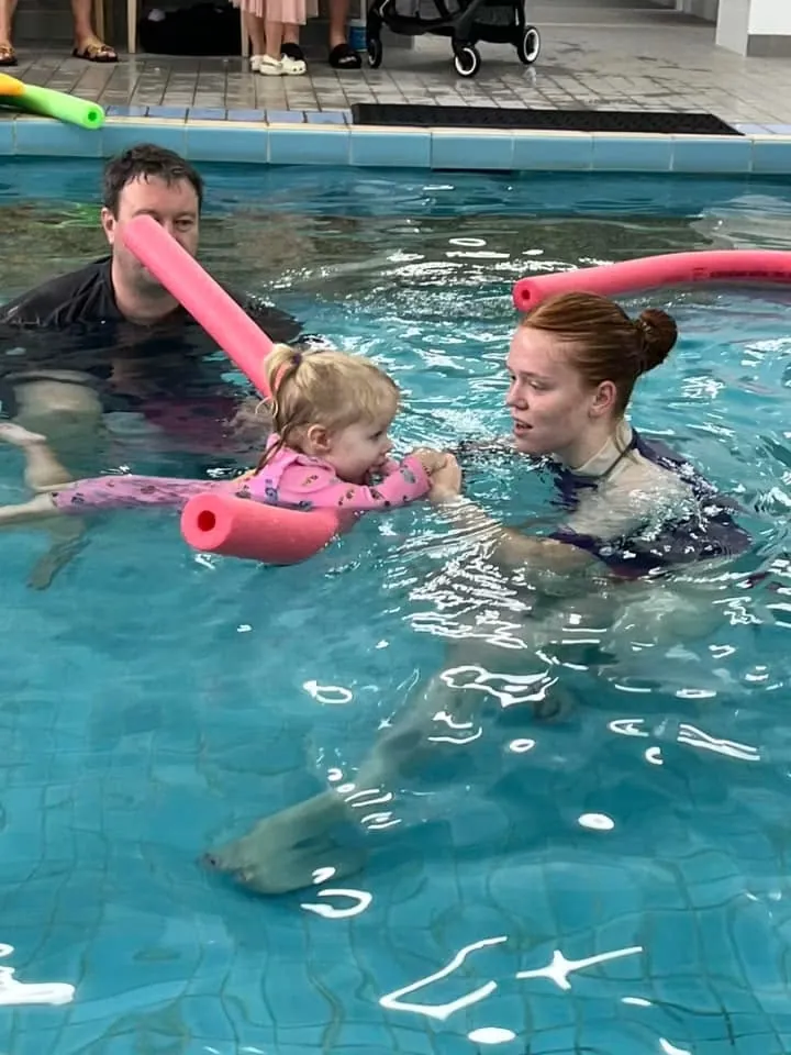 A Pod Squad instructor guiding a toddler on a noodle while a parent watches