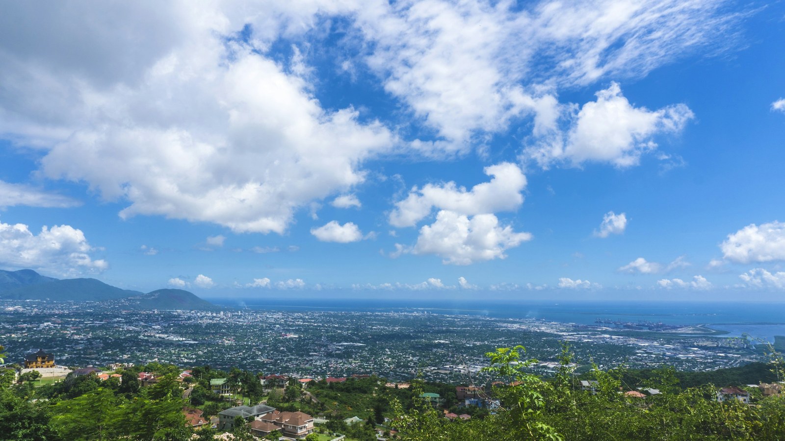 A broad daytime view over Kingston, Jamaica, with dense city buildings below clouds and green hills.