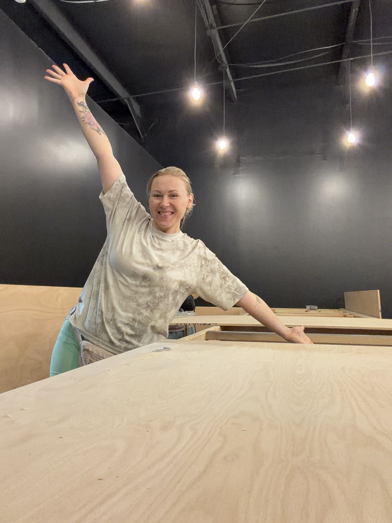 Masha waving with one arm raised, standing behind large plywood panels