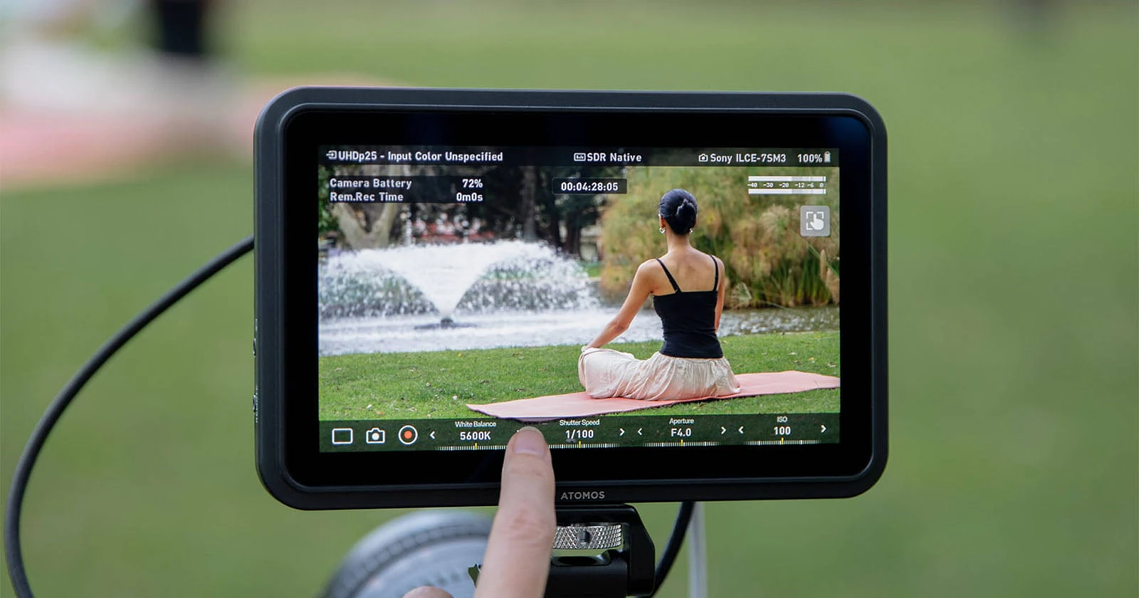 A finger touches the screen of an Atomos monitor displaying a live feed of a woman meditating on a yoga mat in a park.