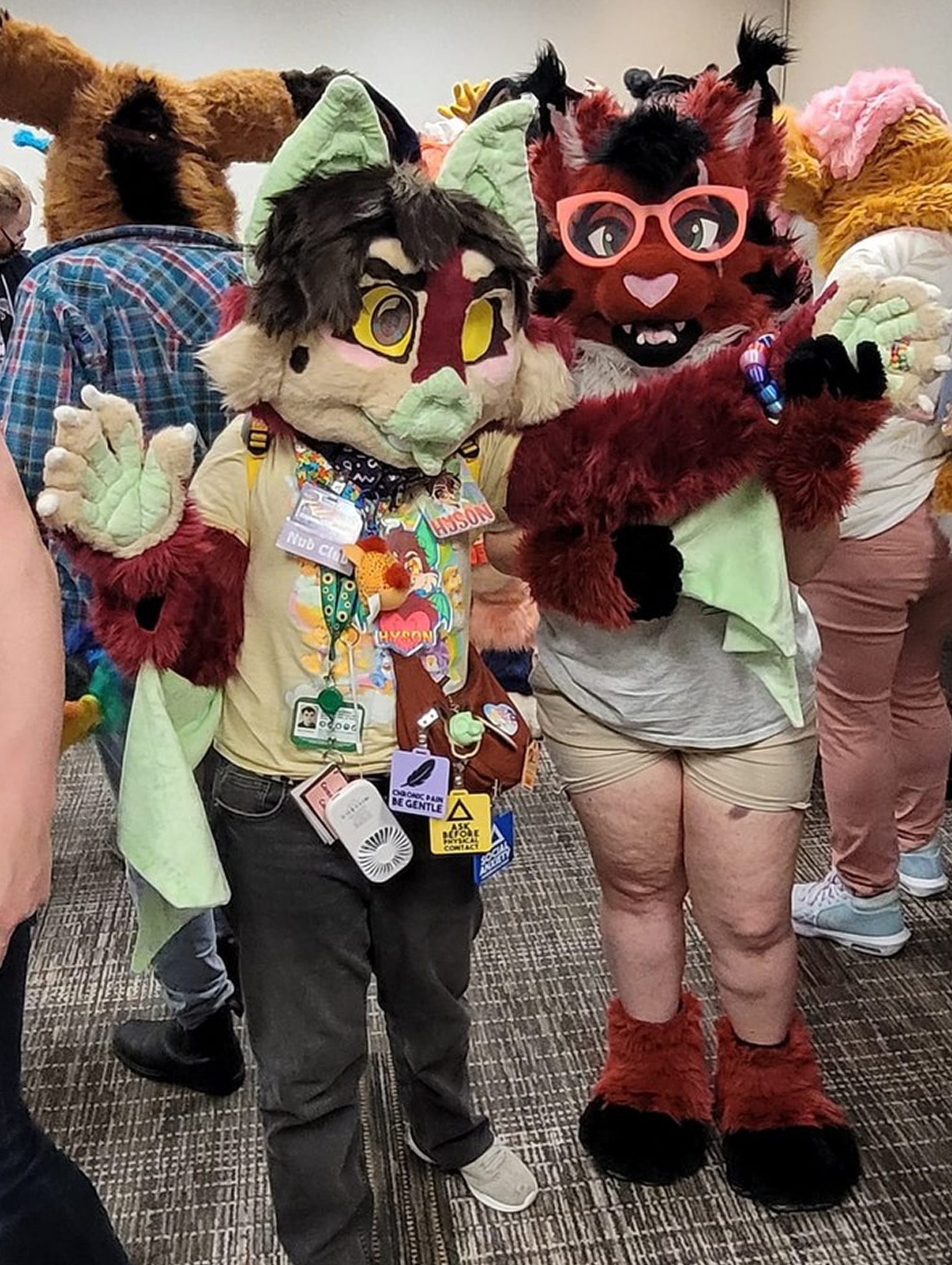 A pair of fursuiters; a multicoloured bat, and a red, white and black lynx, who is holding up one of the bat suiter's arms to show off his wing