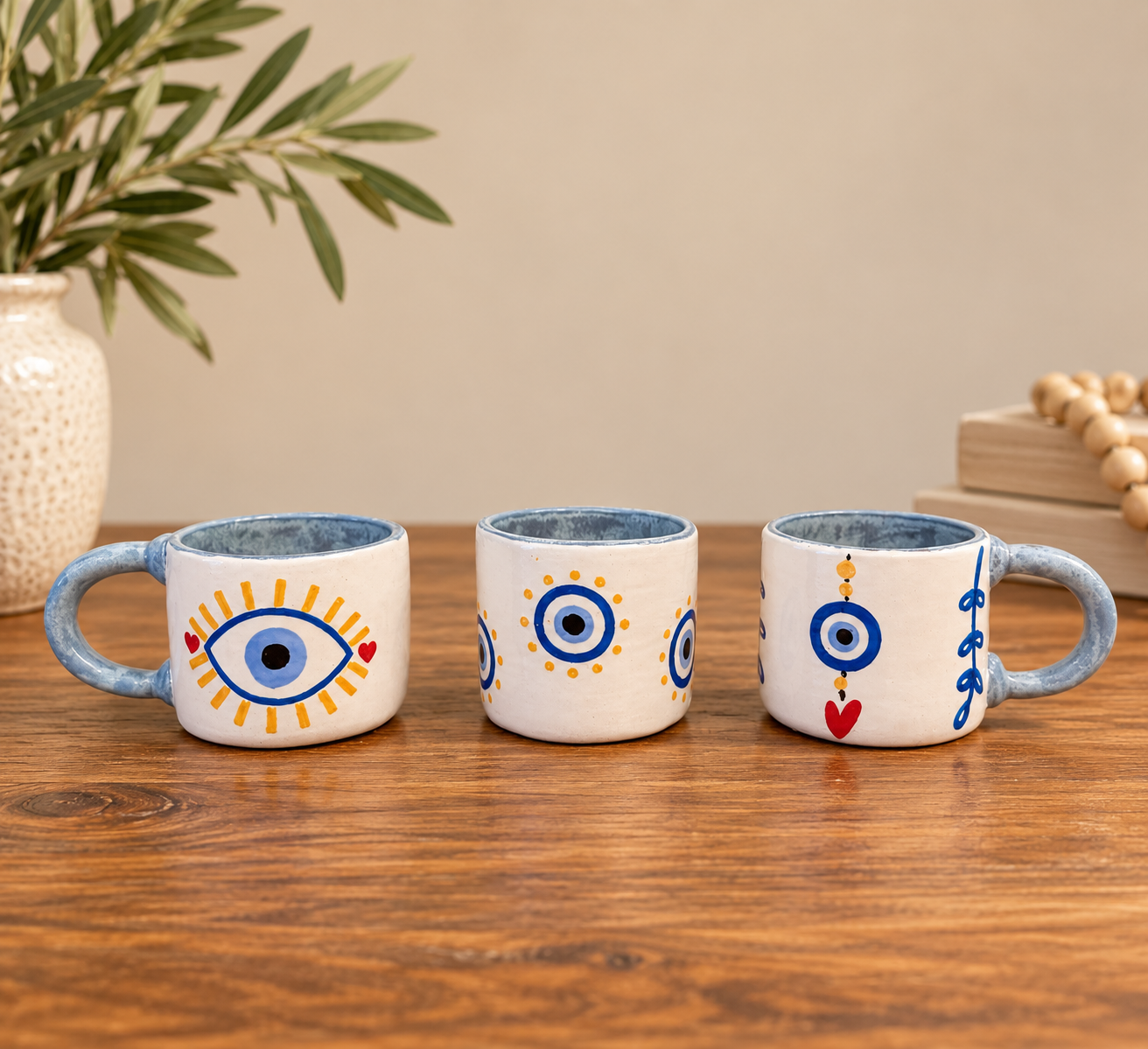 The Evil Eye Espresso Cup Set: three white hand-built cups with light blue handles and interiors, each hand-painted with a different evil eye design, on a wooden table