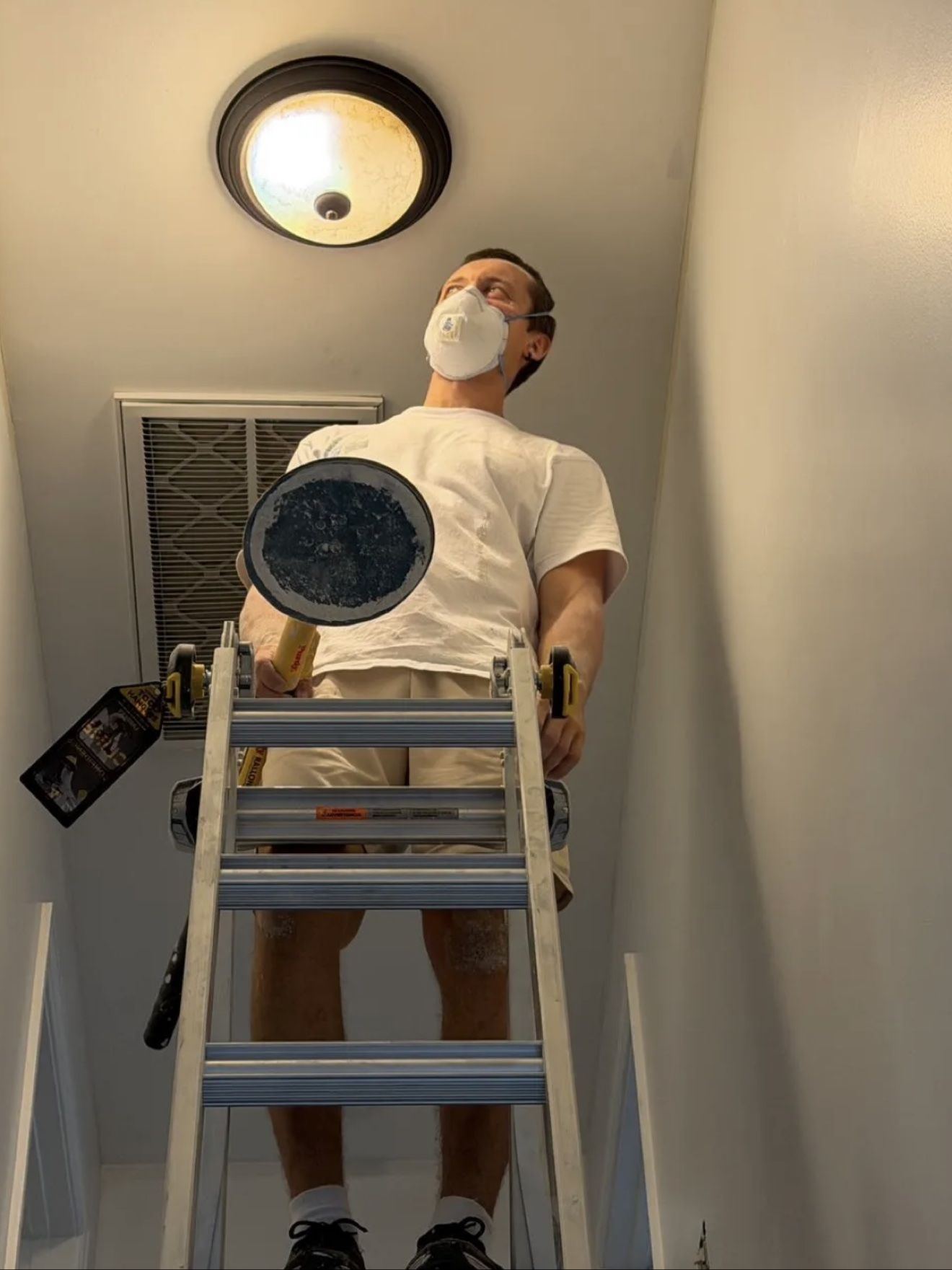 Aleh on a ladder, sanding a ceiling in a respirator mask