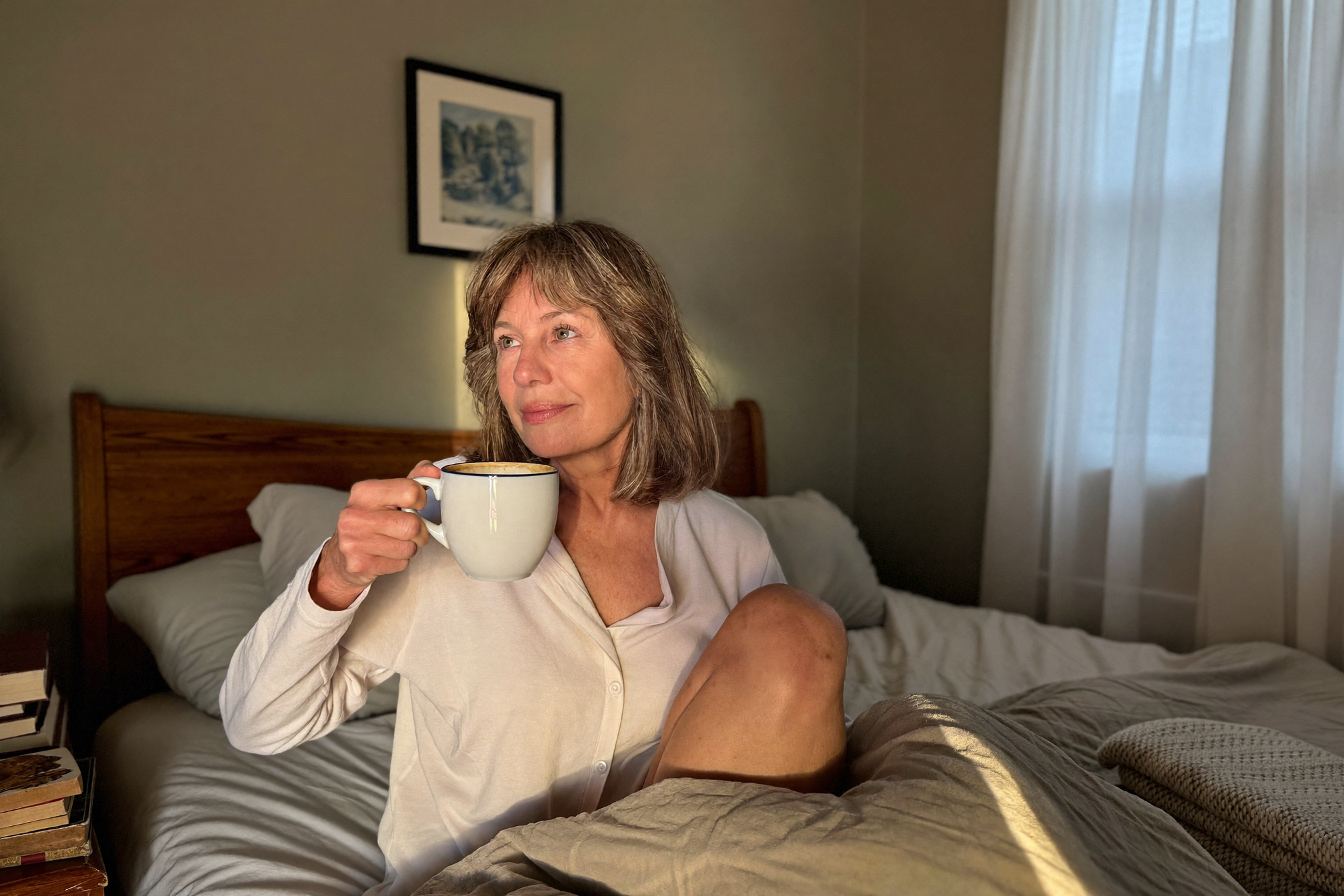A woman waking up peacefully in warm morning light, sitting up in bed with a coffee cup, calm expression.