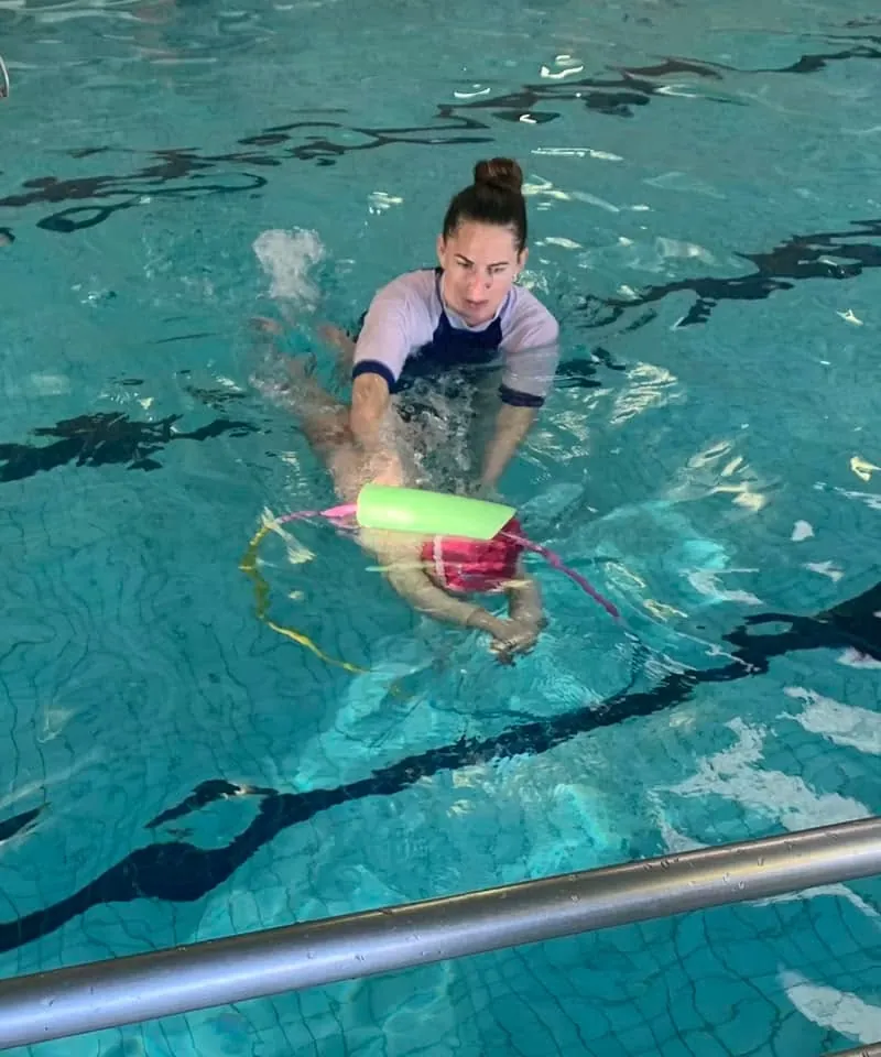 A Pod Squad instructor supporting a child swimming with a pool noodle