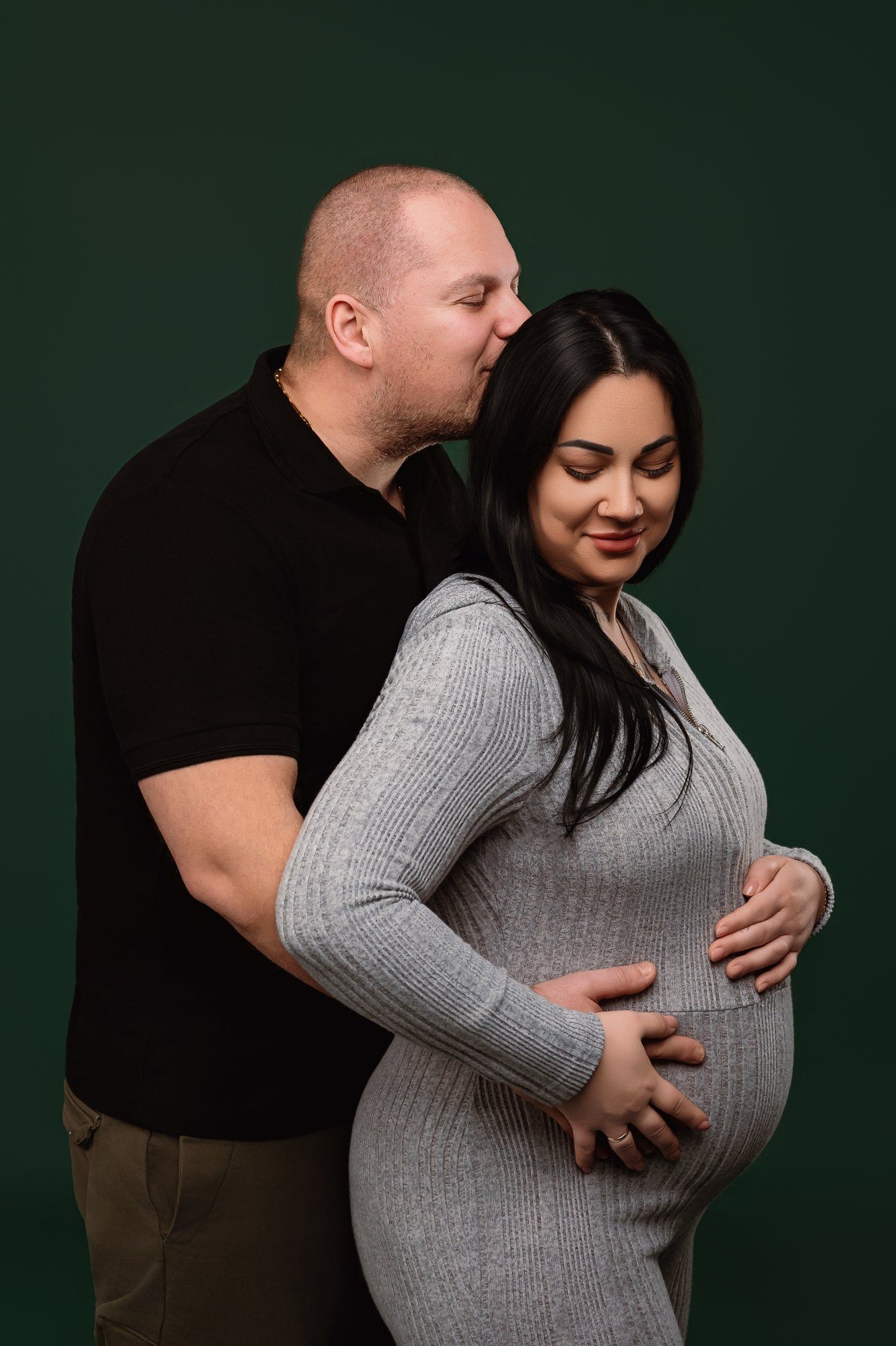 Expecting couple embracing with man kissing woman's forehead against green studio backdrop