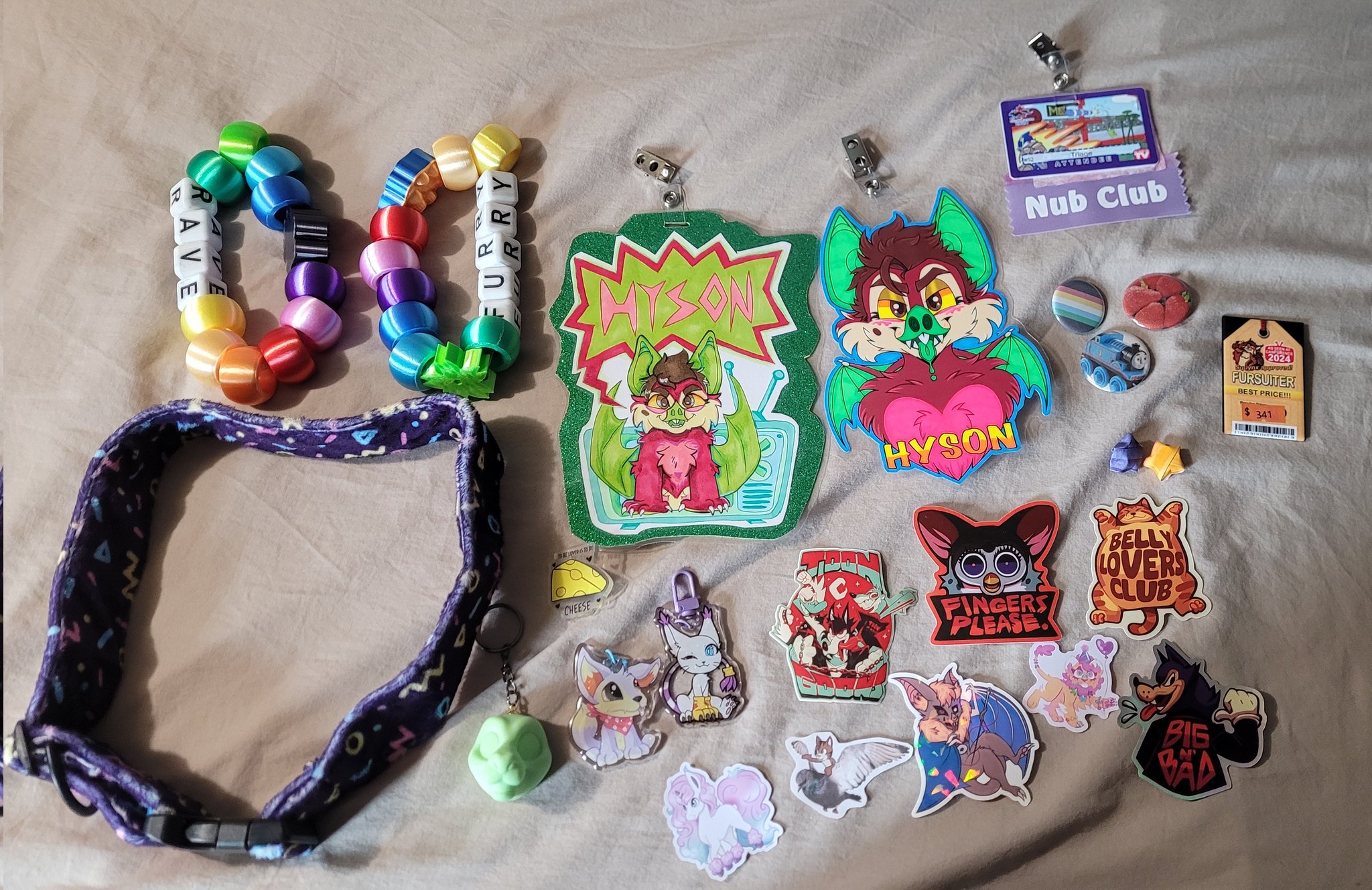 Various items laid out on a bed; two oversized kandi bracelets, a collar, some badges, stickers, keychains, and pins.