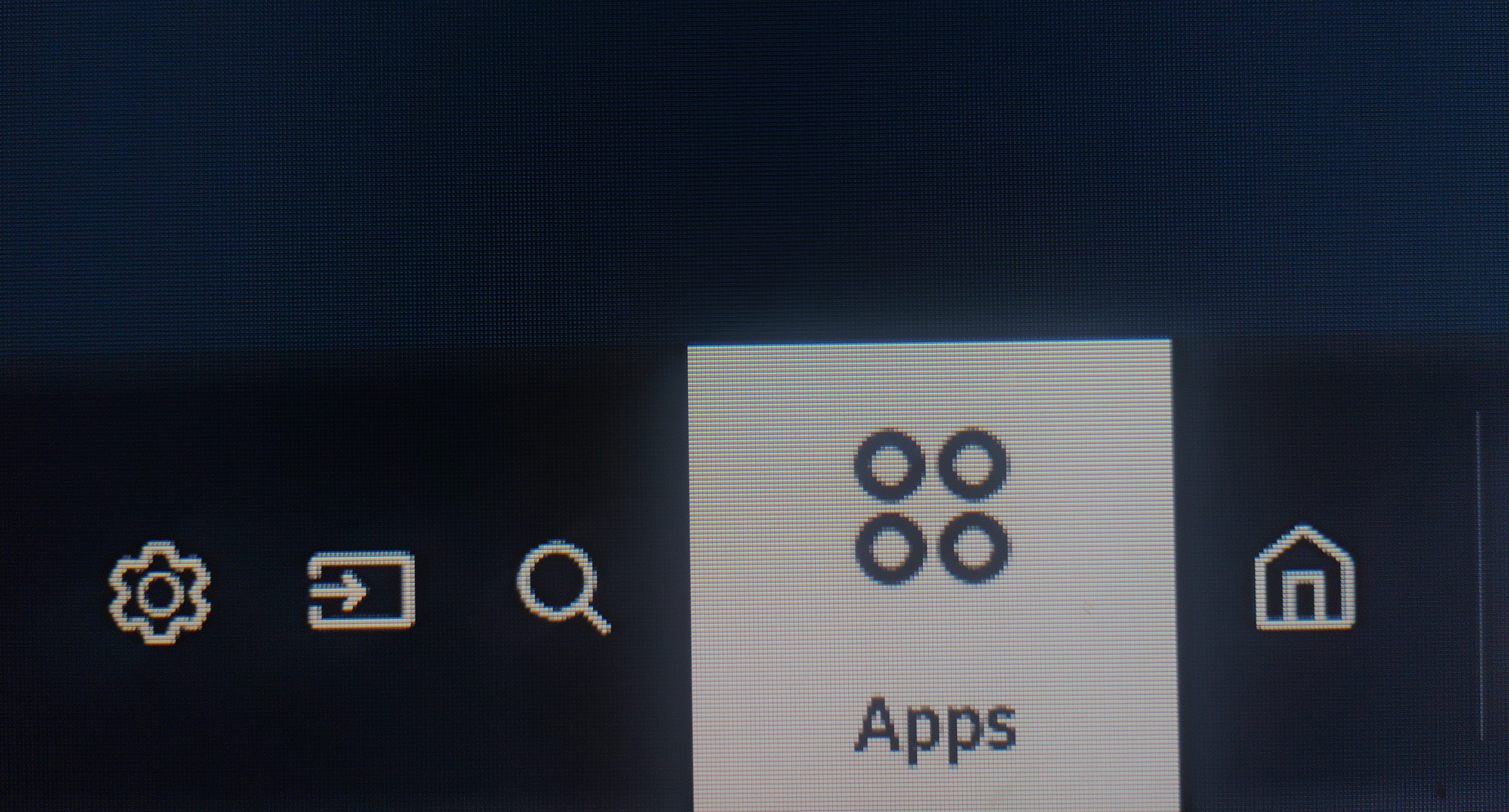 Una interfaz de usuario digital con iconos y texto en una pantalla. En un fondo oscuro, se ven cinco iconos blancos alineados horizontalmente. De izquierda a derecha: un engranaje, una flecha en un rectángulo, una lupa, un cuadrado con cuatro círculos y una casa. El cuadrado central, que contiene el icono de cuatro círculos, está resaltado en un tono claro. Debajo del icono de cuatro círculos, se lee 