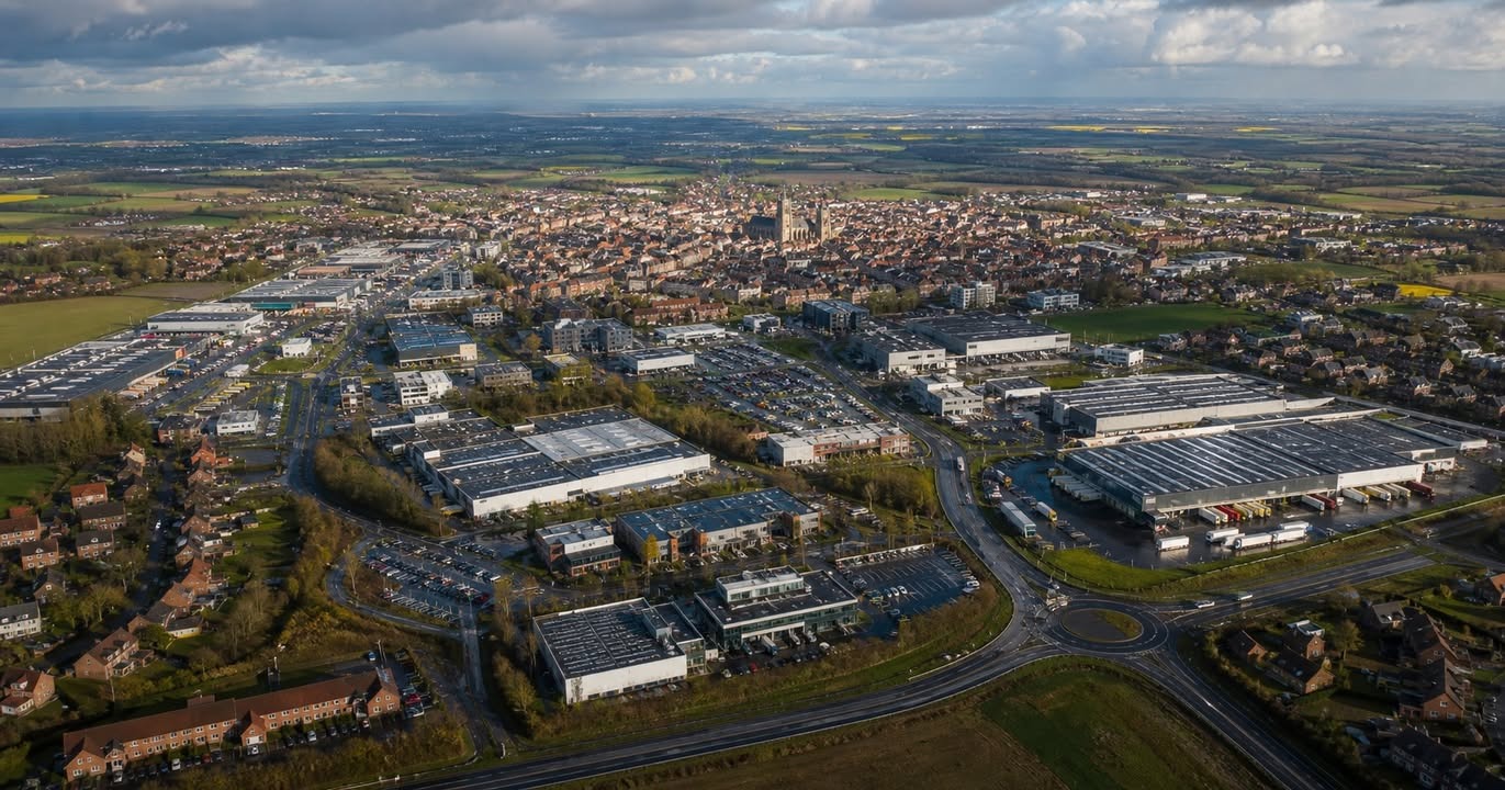 Vue aérienne d'un parc industriel avec de grandes halle et des routes, entouré de zones résidentielles et de champs sous un ciel nuageux.