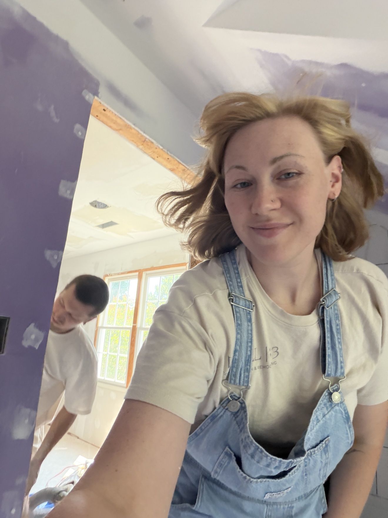 Masha in denim overalls taking a selfie mid-renovation, with Aleh working in the doorway behind her