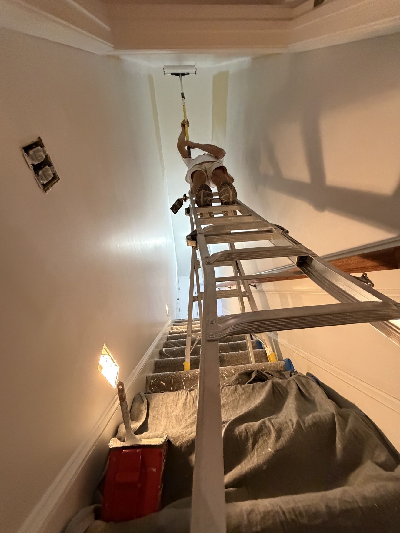 Aleh at the top of a long ladder over a staircase, painting a tall stairwell wall with a roller, seen from below