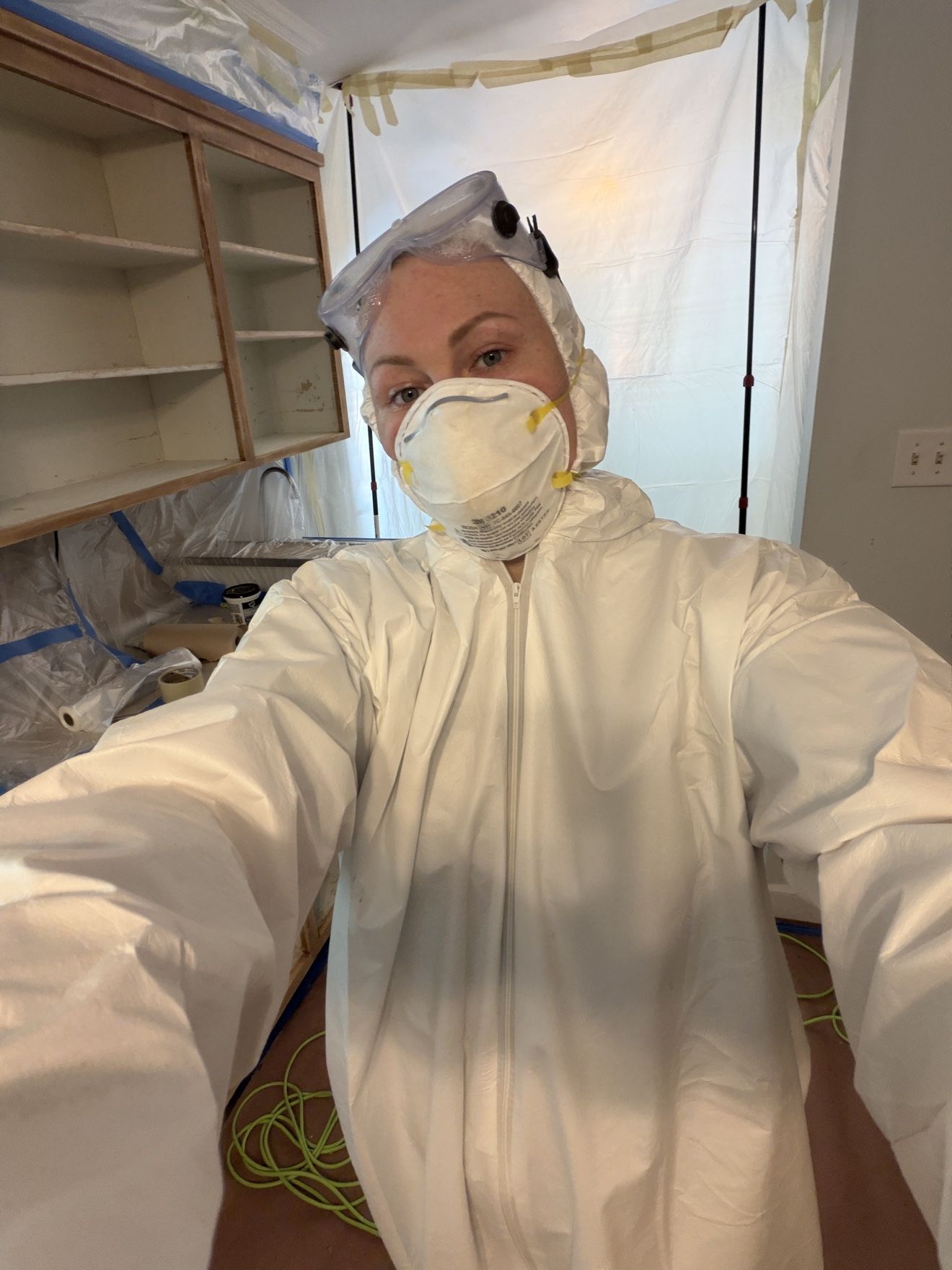 Masha in a white protective suit and respirator mask, taking a selfie in a plastic-covered kitchen
