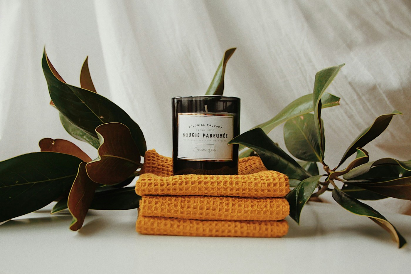 A lit candle resting on folded yellow towels beside a leafy plant and glass bottle.
