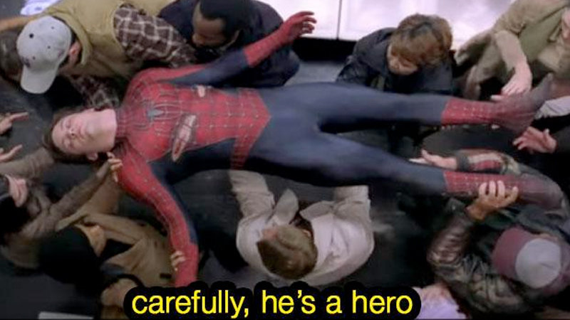 A screenshot from Spiderman 2 wherein an unconscious Spiderman is carried to safety by a crowd of people he protected. The subtitle reads: "Carefully, he's a hero"