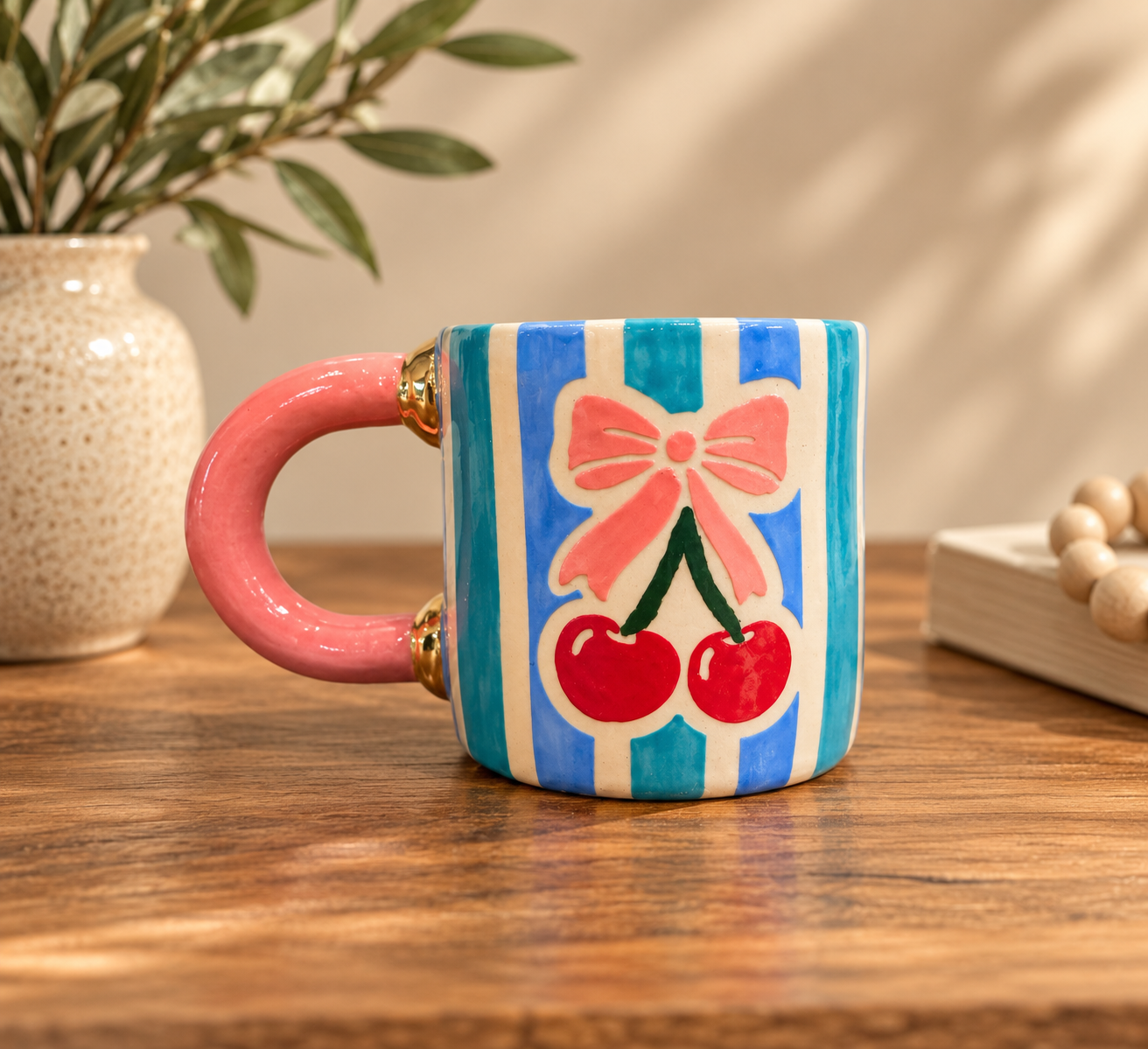 Hand-built mug with blue and teal stripes, hand-painted red cherries with a pink bow, and a pink handle with gold luster