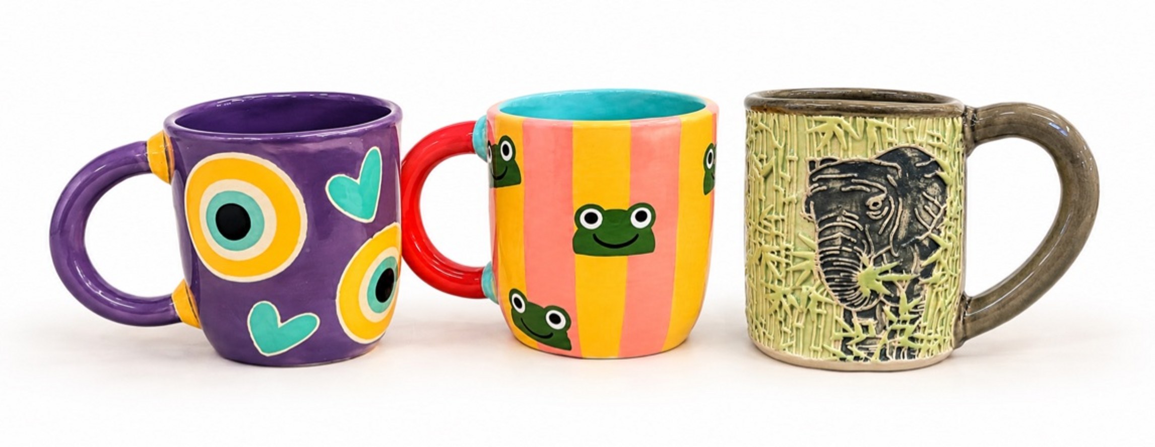 Three of Lisa's handmade mugs on a white background: a purple mug with evil eyes and hearts, a pink-and-yellow striped mug with green frogs, and a green sgraffito mug with an elephant
