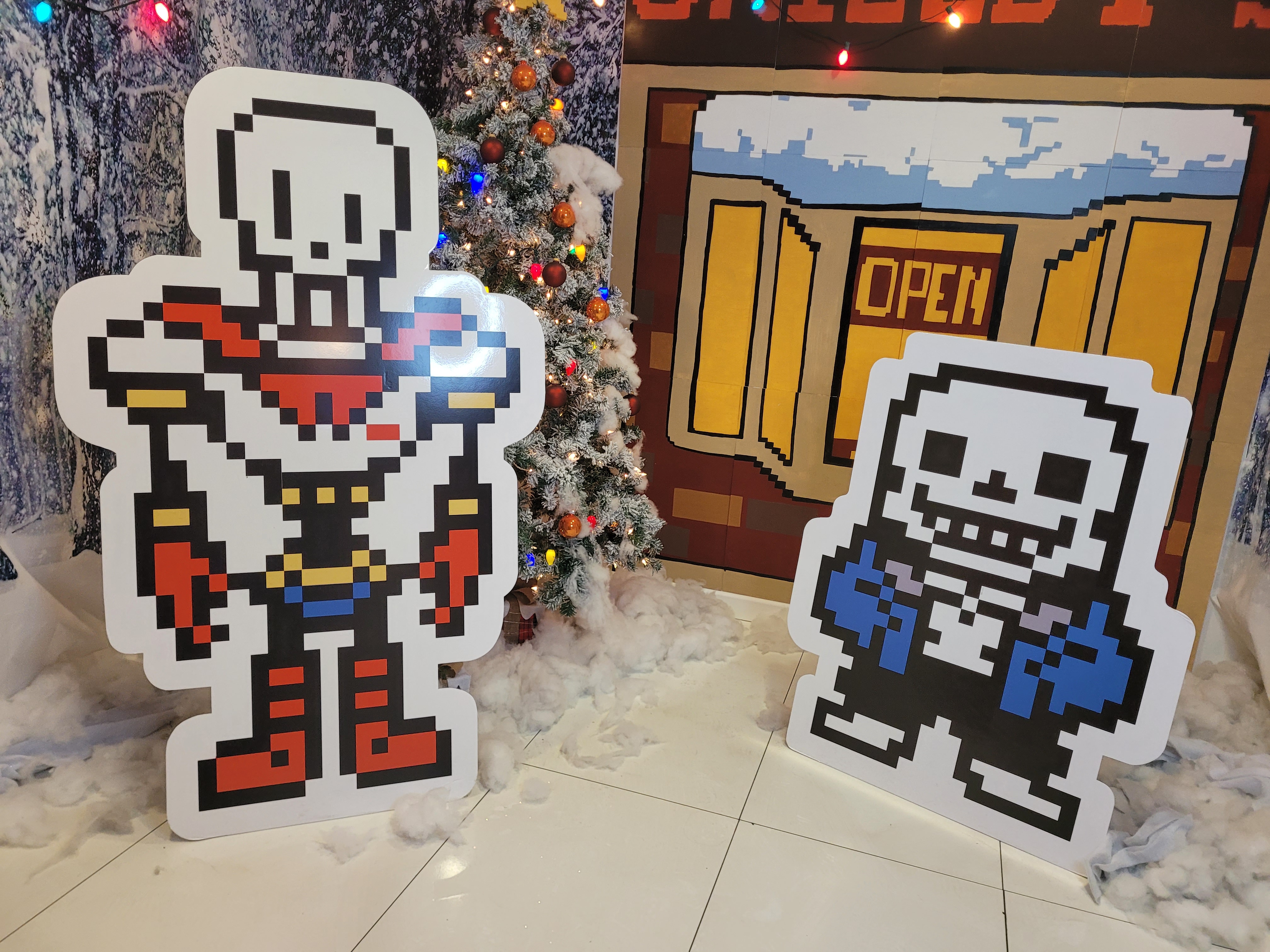 cardboard cutouts of sans and papyrus from undertale. sans is a short skeleton with a wide face, wearing a smile, a blue jacket, black shorts, and slippers. papyrus is a tall skeleton with a long face, and is wearing a red cap, boots, and gloves, as well as blue bottoms. both cutouts are using their less-detailed overworld sprites. they are flanked by a cutout of sans' stand from outside of snowden as well as a christmas tree and a bunch of cotton stuffing, mimicking snow.