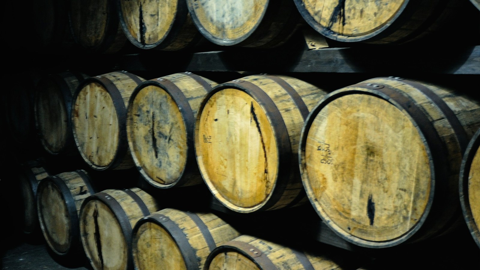 Rows of wooden whiskey barrels stacked in a dim cellar with warm light falling across the oak.