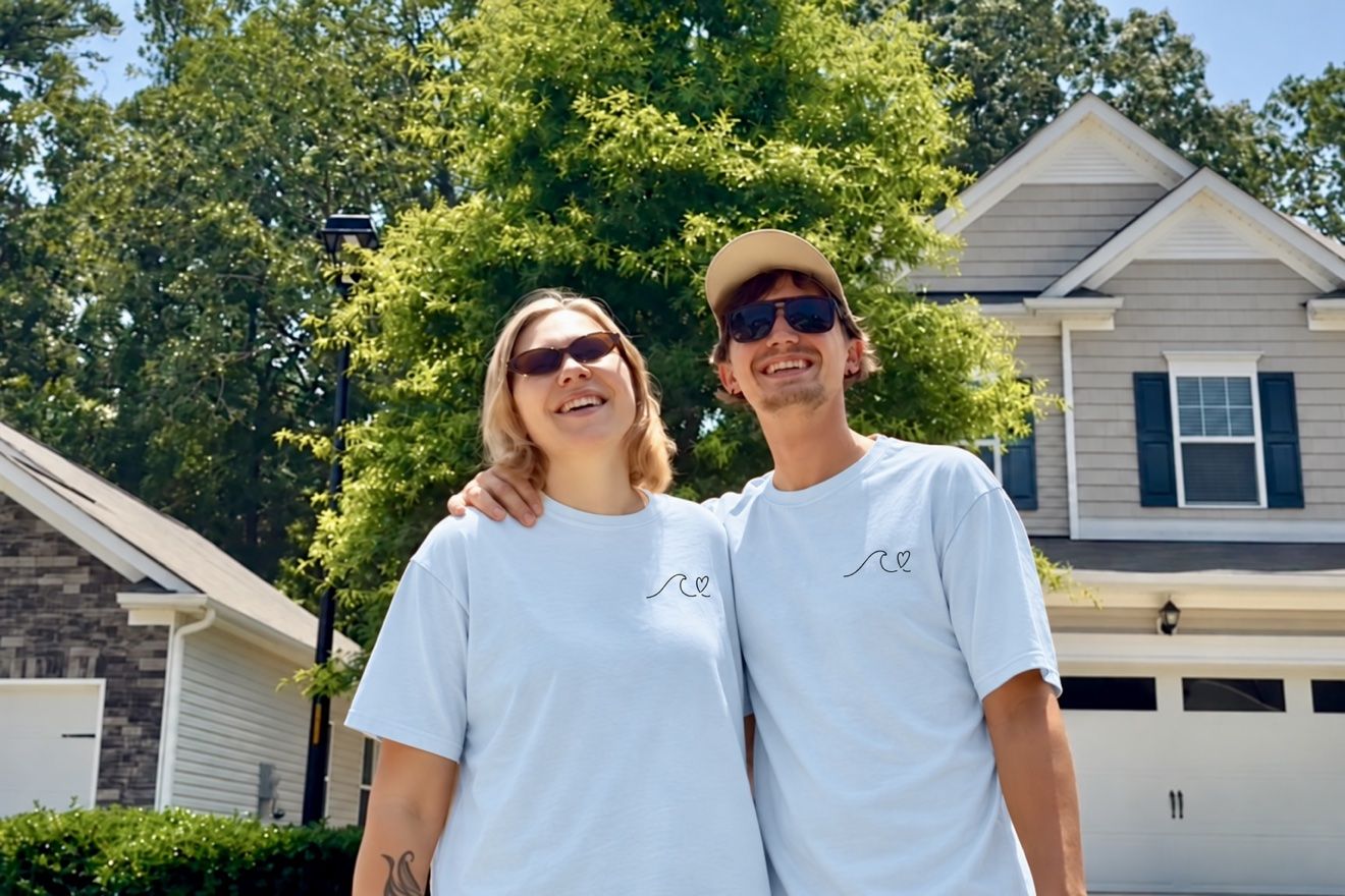 Masha and Aleh in matching light blue t-shirts with a small wave logo, smiling in front of a house with green trees