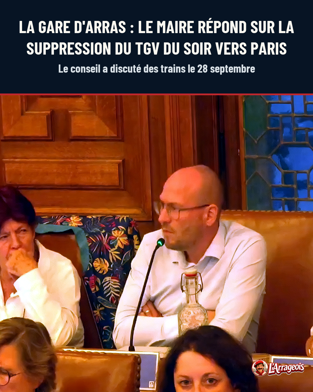 Un homme en chemise blanche parle au micro pendant une réunion, entouré de conseillers dans une salle aux murs en bois.