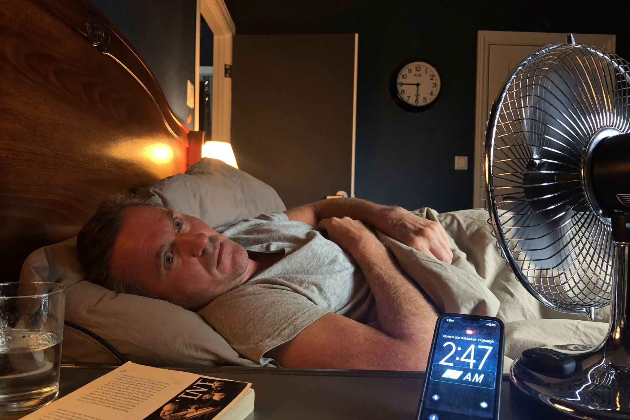 A man in his fifties awake in bed at night, a box fan running loudly on the nightstand.
