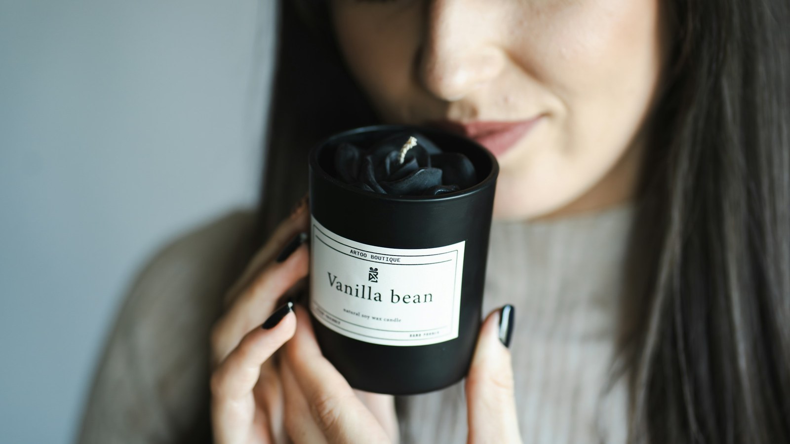 A woman with closed eyes holds a small lit scented candle near her face and smells its fragrance.